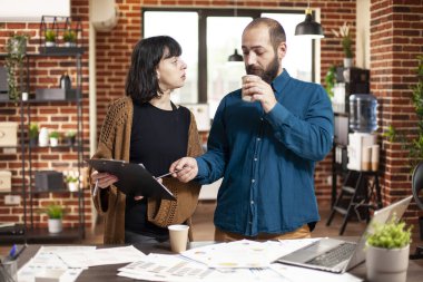 İş adamı kahve içiyor ve yeni başlayan şirketin iş analizlerinde bayan iş arkadaşına yardım ediyor. Elinde dosya olan genç bir kadın, ofis masasındaki sakallı yöneticiye strateji fikirlerini gösteriyor..