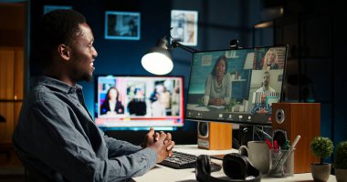 Genç siyahi bir adam bilgisayarında meslektaşlarıyla bir video görüşmesi yapıyor, uzak bir konferansa katılıyor ve ofisinden giyiniyor. Freelancer telekonferansı. Kamera A.