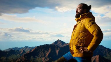 Doğa aşığı nefes kesen bir dağ sırasının kenarında duruyor, yükselen zirvelerin ve uçsuz bucaksız gökyüzünün manzaralı arka planına bakıyor. Macera ve açık hava keşiflerini seven genç bir adam. Kamera A.