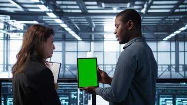 Sunucu odası veri bilimcileri taklit tablet kullanıyor, hataları önlemek için güvenlik denetimleri yapıyor. Veri merkezinde iş arkadaşı olan Afrikalı Amerikalı bir adam yeşil ekran cihazına bakıyor, kamera B