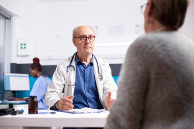 Yaşlı bir erkek doktor, tıbbi randevusu sırasında emekli bir kadınla konuşuyor. Laboratuvar önlüklü kıdemli doktor, klinikteki beyaz kadın hastayla sağlık sonuçlarını ve tedaviyi tartışıyor..