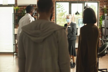 Tuğla duvar ofisinde maske takmış terapi programına hazır bir grup birey bekliyor. Emekli kadın terapist, insanlar odaya girip oturmaya hazırlanırken odanın ortasında bekliyor..