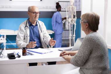 Laboratuvar önlüklü emekli doktor tıbbi konsültasyonda yaşlı bir kadınla konuşuyor. Masasında dosya ve dizüstü bilgisayarı olan kıdemli erkek doktor, beyaz kadın hastaya tedavi planlarını anlatıyor..