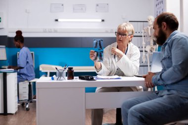 Tıp bürosunda, yetenekli emekli hekim, sakallı erkek hasta konsültasyonu dikkatle dinlerken röntgen görüntüsü tutuyor. Kıdemli kadın doktor klinikteki genç adama radyoloji detaylarını açıklıyor..