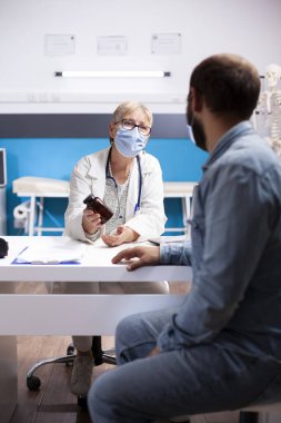 Yaşlı kadın doktor sakallı bir adamla oturuyor ve tedavi için ilaç kullanımını tavsiye ediyor. Kıdemli hekim, erkek hastaya reçeteli ilaç veriyor. İkisi de klinikte maske takıyor..