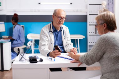 Dost canlısı kıdemli doktor elinde bir şişe reçeteli ilaç tutuyor ve masada oturan emekli kadın hastaya ilaç etkilerini açıklıyor. Hemşire yakınlarda çalıştığı için yaşlı kadınlara danışmanlık yapıyor..