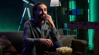 Adam evde kanepede dinleniyor, evde televizyon izlerken ağzına patlamış mısır tıkıyor. Abur cubur yiyen ve neon ışıklı oturma odasında TV şovları yapan kişi, kamera A