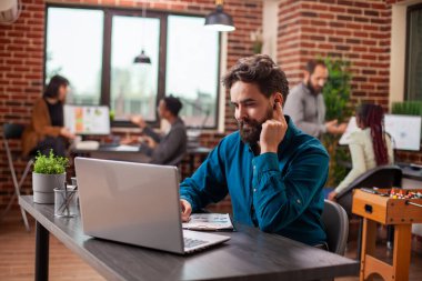 Kablosuz kulaklıklı bir iş adamı, modern işyerindeki bilgisayarında sanal beceri eğitimine katılıyor. Dosyası olan bir erkek ofis çalışanı, kulaklığına dokunuyor ve online sunuma odaklanıyor..