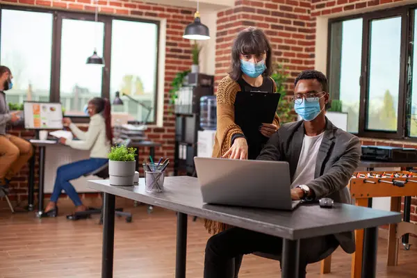 Kadın yönetici durur ve aygıt ekranına işaret eder, erkek iş arkadaşına başlangıç ofisinde veri hatalarını gösterir. Her ikisi de yüz maskesi takan, meslektaşı olarak dizüstü bilgisayarına el kol hareketi yapan beyaz kadın..