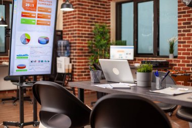 Ekip toplantısına hazırlanmak için dijital cihazlar ve mali belgelerin yer aldığı boş bir toplantı odası. Tuğla duvar iş yeri boş, veri sunumu ve strateji planlaması için monitörler hazır.