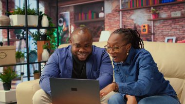 Serbest çalışan çift, yeni bir proje için bilgi toplamak amacıyla evde makaleler okuyarak ve bilgi toplayarak araştırma yapıyor. İnsanlar kanepede birden fazla iş yapıyor ve online raporları inceliyor. Kamera A.