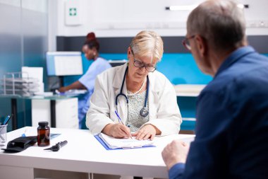 Kıdemli kadın doktor, yaşlı adamın semptomlarını ve klinikteki panoda bulunan belgeleri dikkatle dinler. Emekli erkek hasta için tıbbi talimatlar yazan yaşlı bir doktor..