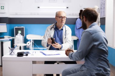 Tıp bürosundaki genç beyaz adam, boynunda steteskop olan yaşlı bir doktor gibi dikkatle oturuyor ve ilacın etkisini anlatıyor. Yaşlı hekim genç hastaya reçeteli ilaç veriyor..