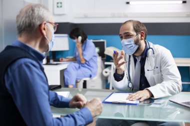 Erkek hekim, emekli hastasıyla oturup sağlık sorunlarını tartışıyor ve sağlık ofisindeki not panosundaki notları inceliyor. Yaşlılara yönelik sağlık tedavi planlarını tanımlayan genç bir sağlık uzmanı.