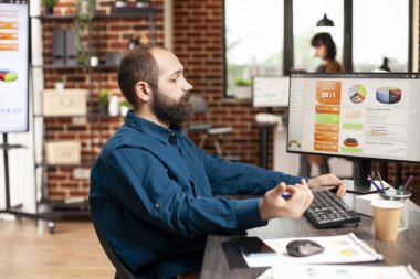 İş yerindeki sakallı adam ofis masasında oturur ve masaüstü monitöründeki mali tabloları analiz eder. Erkek analist, tuğla duvar çalışma alanında yeni başlayan şirket için proje fikirlerini araştırmaya odaklandı.