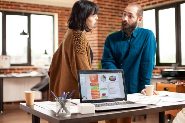 Meslektaşlar tuğla duvar odasında durur, kahve fincanları ve dijital cihazlarla mali evrakları gözden geçirirler. Masadaki dizüstü bilgisayar, iş arkadaşları başlangıç ofisinde iletişim kurarken pazar analizini görüntüler.