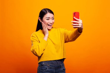 Kazaklı muhteşem Asyalı kadın sarı arka planda cep telefonuyla selfie çekiyor. Genç bayan hareketli bir cihazla kendi portresini çekerken yanağını canlı bir şekilde tutuyor..