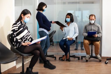 Yüz maskeli siyahi kadın, ofis lobisindeki hamile işverenden teklif alıyor. Muhtemel bayan iş görüşmesi için başvuru toplamaya başladığında çeşitli adaylar evraklarını okuyorlar..