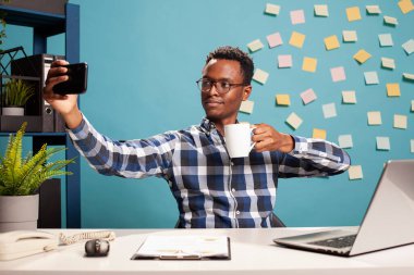 Afrikalı Amerikalı şirket danışmanı selfie çekiyor ve modern çalışma alanında bir fincan çay içiyor. Cep telefonlu siyah bir işadamı masasında kahve içerken çekilmiş bir videosunu çekiyor..