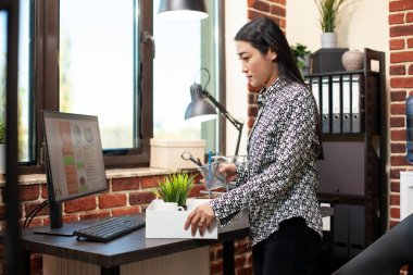 Asyalı profesyonel masa başında duruyor, tuğladan duvar işyerindeki bir fabrika da dahil olmak üzere ofis kağıtlarını düzenliyor. İş kadını, başlangıçta iş eşyalarını beyaz kutuya koyarak ayrılmaya hazırlanıyor.