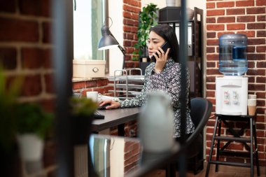 Kadın girişimci bir müşteri konuşması için akıllı telefonunu kullanıyor ve masaüstü monitörüne bakıyor. Asyalı yönetici, bilgisayardan güncellemeleri referans alan mobil cihazıyla iş arkadaşıyla konuşuyor.