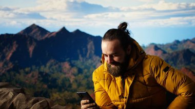 Gezgin oturuyor ve telefonunu kayalık dağın tepesinde karıştırıyor, çarpıcı dağ sırasından ve manzaradan memnun. Teknoloji ve internete kafayı takmış. Kamera A.