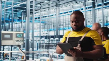 Akıllı fabrika mühendisi tesis katındaki robot silah ve montaj hatlarını denetliyor. Endüstri 4.0 Afro-Amerikan erkeği sistem performansını izliyor ve sorun yaratıyor, kamera A