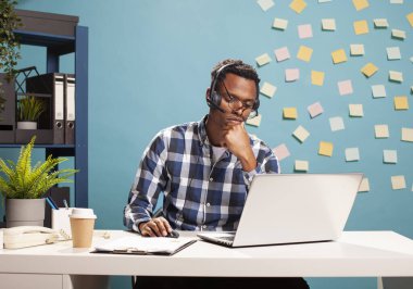 Çok çalışan bir danışman, yeni başlayan bir proje için çözümler üretiyor. Eğri gözlükleri ve kulaklıkları olan siyah bir iş adamı laptopunu kullanarak görevleri çözmeye çalışıyor..