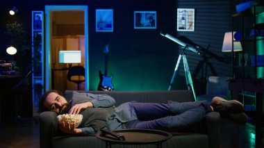 Evdeki bunalımlı adam kendini kötü hissederek kanepede uzanıp televizyon seyrederek ve patlamış mısır yiyerek teselli arıyor. Somurtkan insan depresyonda yatıyor, daha iyi hissetmek için televizyon izliyor, kamera.