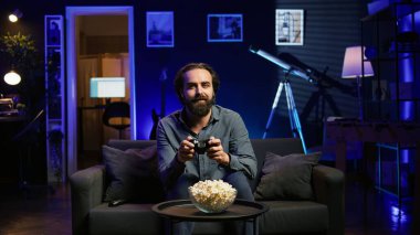 Heyecanlı adam neon ışıklı dairede elinde kontrolörle video oyunları oynuyor. Konsol sistemindeki PVP oyununa katılmak için joystick kullanan mutlu oyuncu, rahatlıyor ve eğleniyor, kamera B