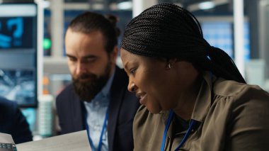 Fabrika müdürü, otomasyon teknolojisini endüstriyel fabrikaya entegre etmek için çalışıyor, cihaza bakıyor. Afro-Amerikalı kadın iş modeli verilerini tablet üzerinde analiz ediyor, üretimi genişletiyor, kamera B