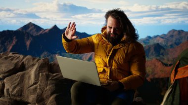 Dağ maceracısı, aşırı rüzgara rağmen dizüstü bilgisayarı üzerinde çalışarak çevrimiçi işini güvenlikten üstün tutuyor. Nefes kesen manzara, onun internet bağımlılığı tarafından gölgeleniyor. Kamera A.