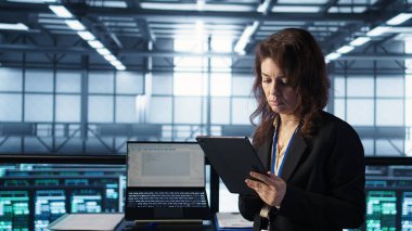 Yeni sunucu mimarileri yerleştirmek için tablet üzerinde yapay zeka teknolojisi kullanan veri merkezinde yönetici. Sunucu odasındaki kadın sabit performansı korumak için depolama dizilerini güncelliyor, kamera B