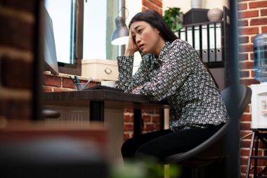 Bilgisayarla masasında oturan yorgun bayan çalışan, başlangıç ofisindeki mali işlerden dolayı yorgun görünüyor. Asyalı girişimci avucunun içinde dinleniyor ve modern çalışma alanındaki şirket iş yükünü inceliyor..