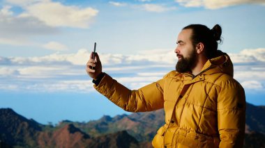 Erkek içerik yaratıcısı bulutlardaki dağ zirveleriyle çevrili bir video kaydediyor, bir dağ tepesine tırmanıyor ve sosyal medya izleyicileri için canlı yayın yapıyor. Maceracı ruh. Kamera B.