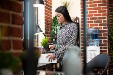 Tuğla duvar çalışma alanında girişimci, duruyor ve beyaz bir kutudan onun saksı bitki ve eşyalarını açıyor, iş günü için hazırlanıyor. Asyalı bir çalışan başlangıç ofisinde masasını kuruyor..