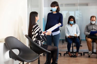 Lobide oturan Asyalı kadın, işe alım toplantısına hazır hamile alıcıya özgeçmiş veriyor. İş başvurusu yapan kadın ve beyaz kadın panoyu tutuyor, görüşme sürecini tartışıyorlar..