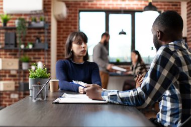Profesyonel erkek istihdam memuru, tuğla duvar çalışma alanında yapılan bir iş toplantısında kadın adayları değerlendirirken görülüyor. Afrika kökenli Amerikalı insan kaynakları müdürü elinde bir dosya tutuyor ve beyaz bir kadınla röportaj yapıyor..