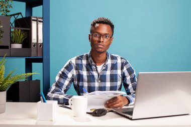 Gözlüklü Afro-Amerikan bir adam masada oturmuş, belgeler ve dizüstü bilgisayarla araştırma yapıyor. Elinde kağıtlarla kameraya bakan siyah bir girişimcinin portresi..