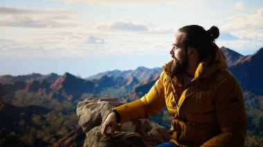 Kaşif, bir dağ zirvesine ulaşmanın heyecanını kucaklıyor ve dinleniyor, temiz havayı ve panoramik güzelliği hissediyor. Yürüyüşçü heyecanlı ve yorgun, kendini geliştirme ve hedefleriyle gurur duyuyor. Kamera A.