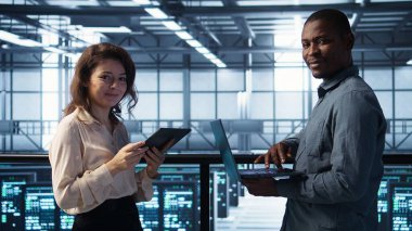 Veri merkezinde dizüstü bilgisayar kullanan gülümseyen yöneticiler, sunucu viralizasyonunu yönetiyorlar. Neşeli bilişim uzmanı ve sunucu odasında not defteri kullanan Afro-Amerikan bir adam, altyapı güvenilirliğini sağlamak için, kamera B