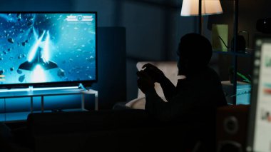 Genç yetişkin, akıllı televizyonunda video oyunlarıyla eğleniyor. Oturma odasındaki LCD monitörüne konsolunu bağlıyor ve evde oyun gecesinin tadını çıkarıyor. Afrikalı Amerikalı oyuncu. Kamera B.