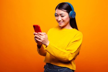Neşeli genç bir kadın, mavi kulaklıkla müzik dinlemekten ve cep telefonuyla mesajlaşmaktan hoşlanıyor. Sarı kazak giymiş Asyalı bir kadın, cep telefonuna gülümsüyor, izole edilmiş bir arkaplanda görüntülü konuşma yapıyor.