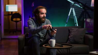 Neon ışıklı evde video oyunu oynayan odaklanmış bir adam, elinde kontrol cihazı tutuyor. Oyuncu büyüleyici bir oyuna bağımlı, çevrimiçi turnuvaya katılmak için heyecanlı, diğer oyuncularla mücadele ediyor, kamera A