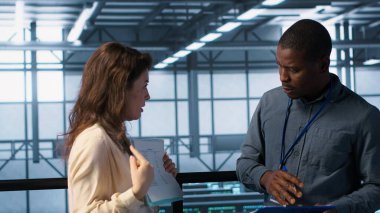 Bilgi işlem uzmanlarından oluşan bir ekip veri merkezi kontrol listesi evraklarını inceliyor. Afrikalı Amerikalı adam ve iş arkadaşı sunucu çiftliğinde işbirliği yapıyor. Belge dosyaları, kamera A