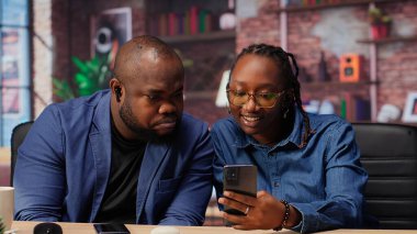 Genç siyahi çift kulaklıklarını paylaşıp akıllı telefondan Webinar dinliyor ve evdeki serbest işlerinde birlikte çalışıyorlar. Erkek ve kadın bir iş projesi hakkında tartışıyorlar. Kamera B.