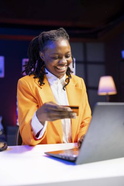 İnternet alışverişi yapan gülümseyen genç bir yetişkin dairede dizüstü bilgisayarına kredi kartı bilgilerini dikkatlice yazıyor. Neşeli Afro-Amerikalı kadın oturma odasında bankamatik kartıyla alışveriş yapıyor.