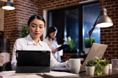 Genç Asyalı girişimci, rahat, modern bir ofiste uzaktan çalışıyor, bir masada oturuyor, tablet ve dizüstü bilgisayar kullanıyor. Gece atmosferi ve açık tuğla duvar çalışma alanı estetiğini geliştirir..