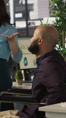 Dikey video çalışanları ve engelli iş arkadaşları dahil ofislerdeki gelir rakamlarına bakıyorlar. Tekerlekli sandalyedeki adam iş arkadaşlarıyla çalışıyor, şirket istatistiklerini karşılaştırıyor.