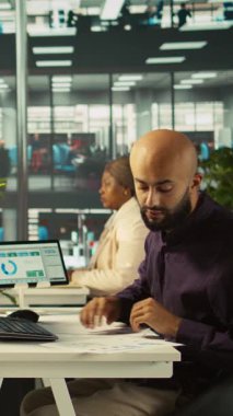 Ofis masasındaki masaüstü bilgisayarındaki belgeleri düzenleyen dikey video çalışanı, raporlar ve dosyalar güncelleniyor. İşçi masadaki bilgisayar ekranında veri grafiklerini analiz ediyor, istatistiksel bilgileri yorumluyor